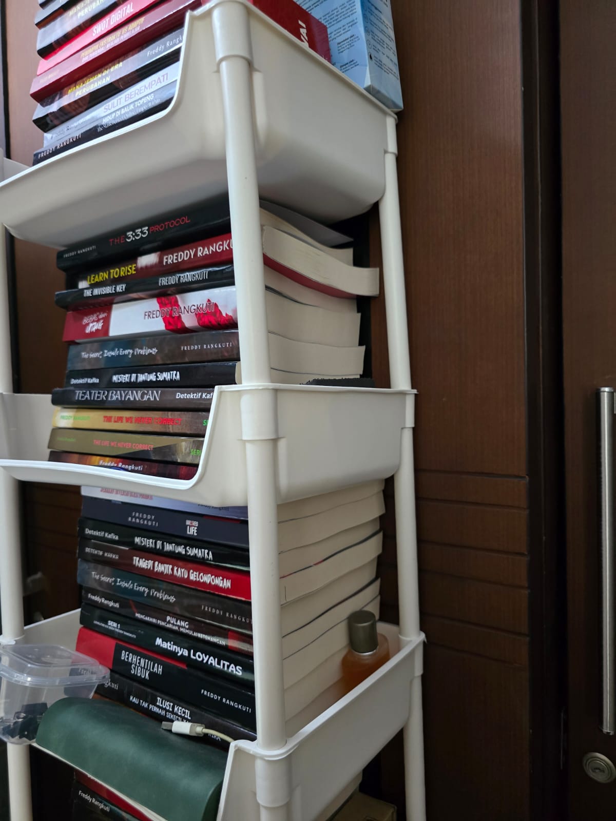 [FOTO 2: Tumpukan buku batch pertama di rak di atas meja] (Dua tahun kerja keras akhirnya menjadi sesuatu yang bisa disentuh).